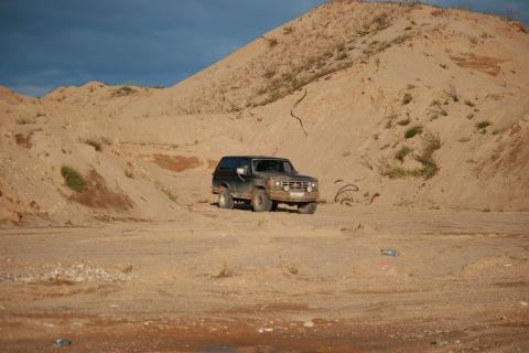 1987 Ford Bronco - 1987 Ford Bronco navigating a sandy terrain under a partly cloudy sky.