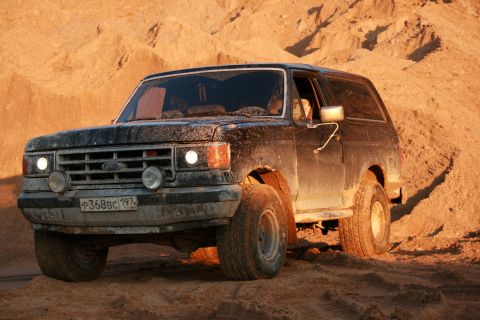 1987 Ford Bronco - A black 1987 Ford Bronco navigates a sandy terrain under warm lighting.