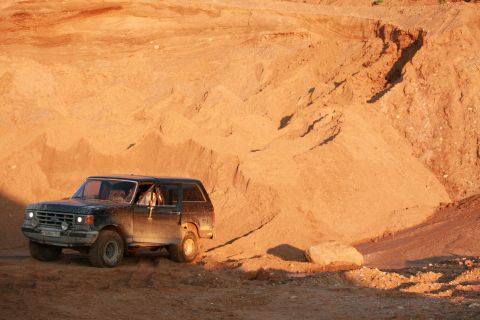 1987 Ford Bronco - A dark 1987 Ford Bronco navigates a sandy terrain, with its doors open.