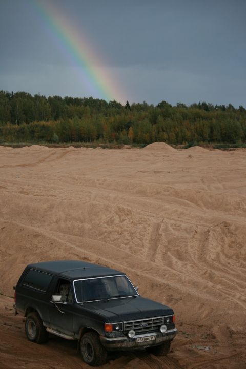 1987 Ford Bronco - A dark SUV sits on sandy terrain under a cloudy sky with a rainbow arching overhead.