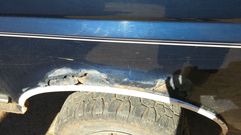 Rust damage and peeling paint above rear wheel arch trim on dark blue vehicle with off-road tire