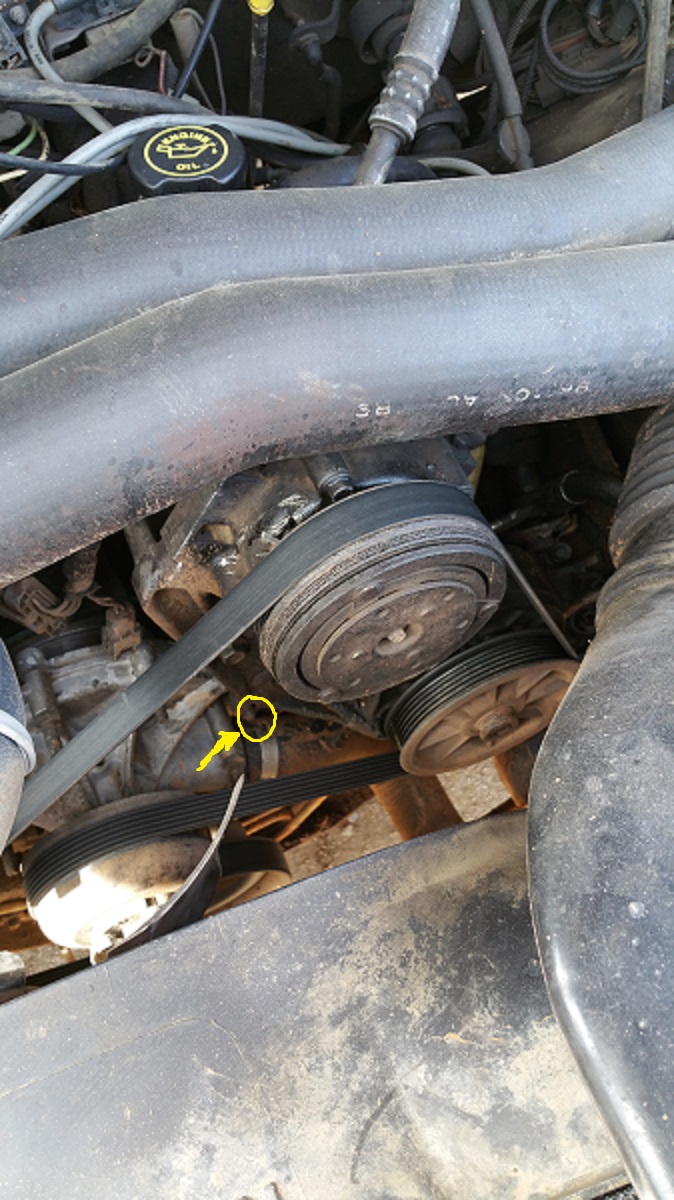 Engine bay showing serpentine belt system, pulleys, and AC compressor. Yellow arrow points to a bolt
