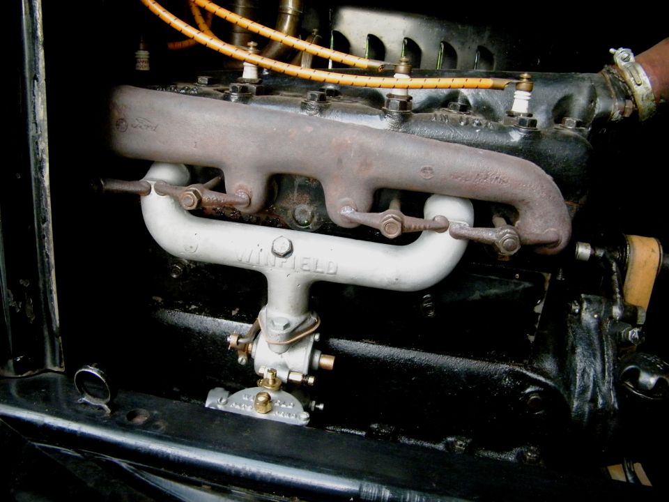 Close-up of an engine with a Winfield carburetor and exhaust manifold. Spark plug wires are visible