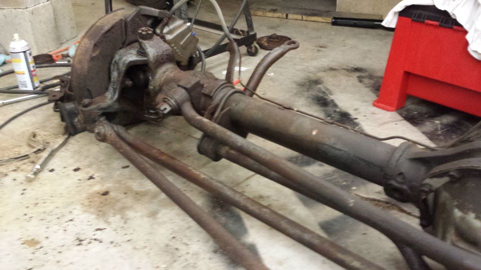 Rusty front axle assembly with steering linkage and brake components, lying on a concrete floor