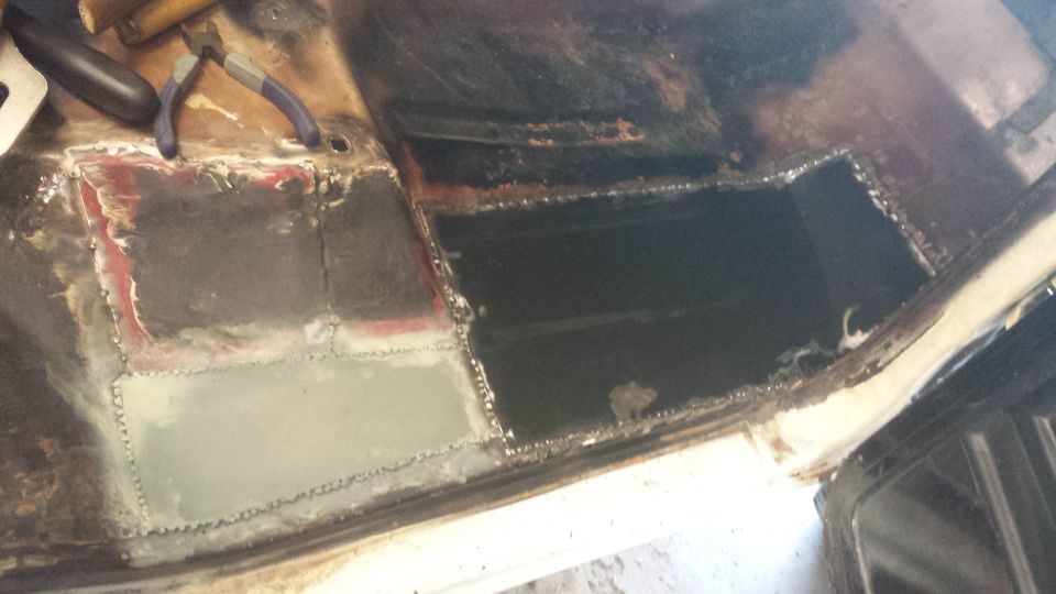 Vehicle floor pan with cut-out section and welded patches, showing rust repair. Pliers rest on top
