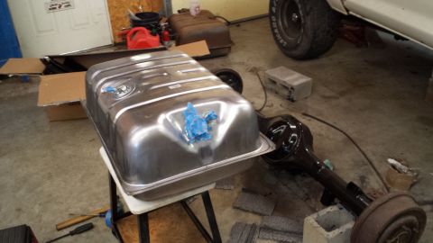 1978 Ford Bronco - A shiny new fuel tank sits on a stand next to a rear axle, with a partial view of a vehicle's tire and fender in the background.
