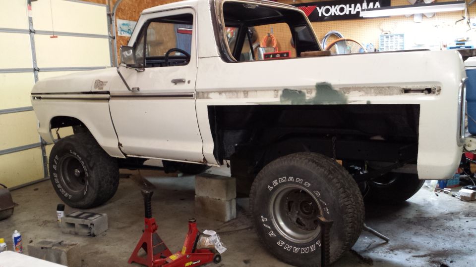 White 1978 Ford Bronco on jack stands in a garage, undergoing repairs. The rear fender area is missing bodywork
