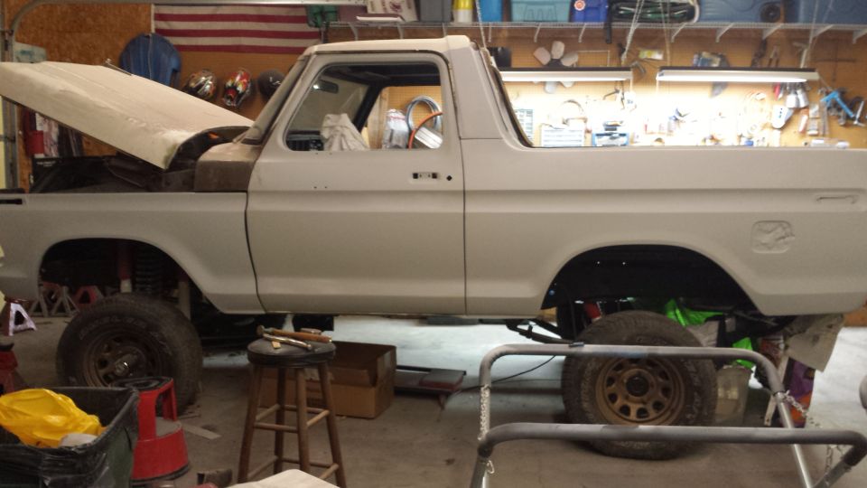 1978 Ford Bronco in primer, hood open, on jack stands in a garage