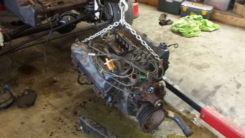Engine suspended by a chain hoist, with vehicle frame and suspension visible in background