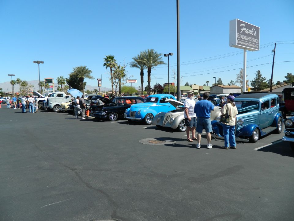 Several classic cars parked in a lot under a bright blue sky, with people milling around