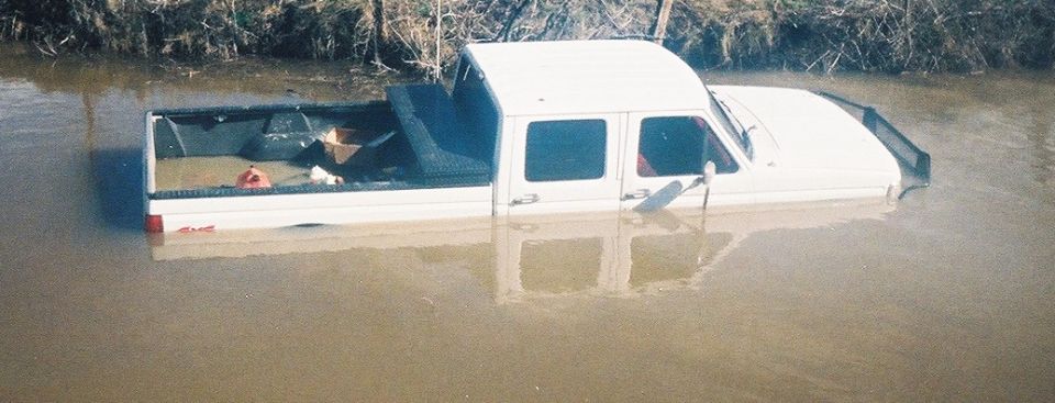 White crew cab pickup truck submerged in muddy floodwater, debris in bed