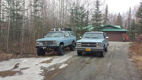 1990 Chevrolet Suburban - Two older model Chevrolet Suburbans are parked outside a house with a wooded background.