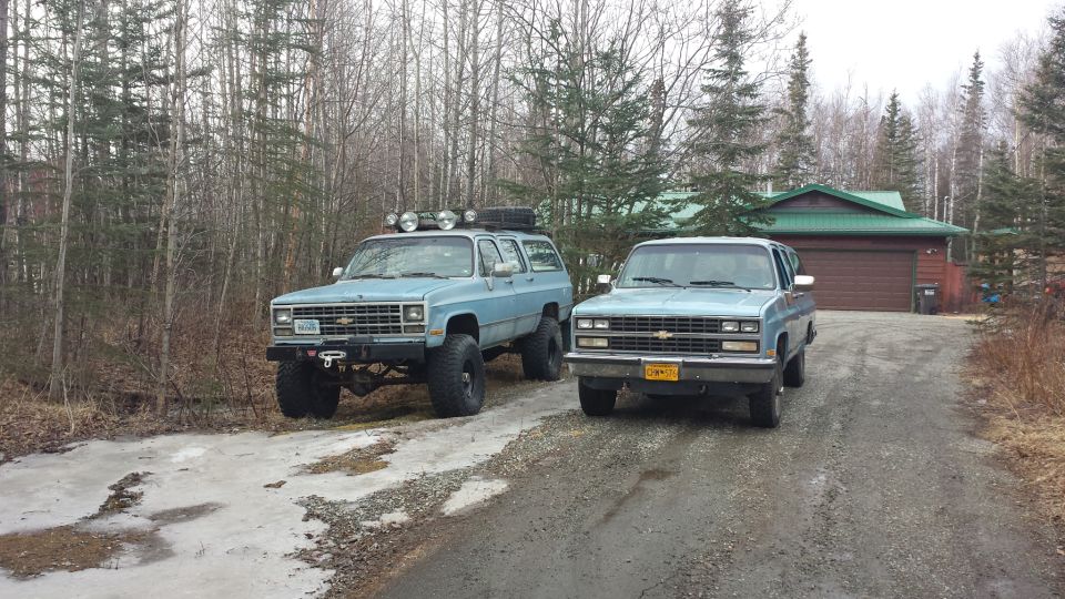 Two light blue Chevrolet Suburbans parked on a gravel driveway in front of a house