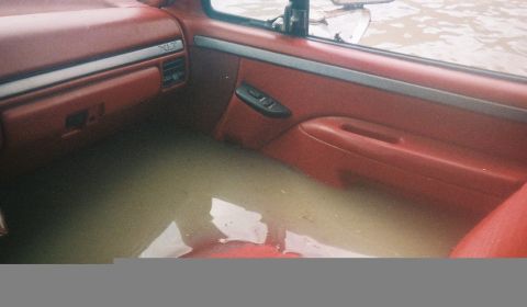 1991 Ford Bronco - Interior of a red-upholstered Ford truck/SUV cabin flooded with murky brown water reaching up to the seats, showing the passenger door pane…