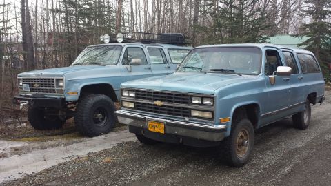 1990 Chevrolet Suburban - Two light blue Chevrolet Suburbans are parked on a dirt road, with trees in the background.