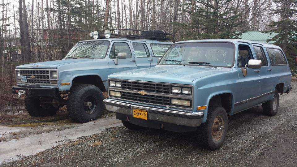 Two light blue Chevrolet Suburbans parked side-by-side, one with off-road modifications
