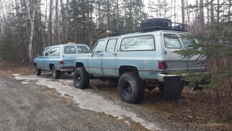 1990 Chevrolet Suburban - Two light blue Chevrolet Suburbans, one modified with off-road tires and a roof rack, are parked on a gravel road.