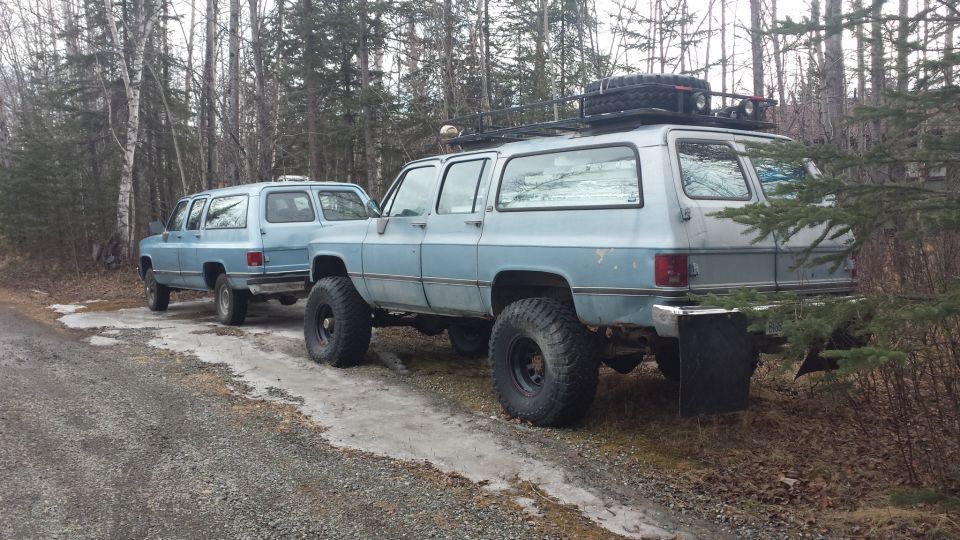 Two light blue 1990 Chevrolet Suburbans with off-road tires parked side-by-side