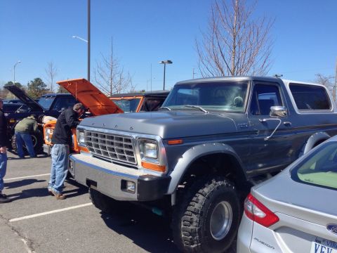 1979 Ford Bronco - Several vehicles are parked in a lot, with people gathered around the open hood of an orange Bronco.