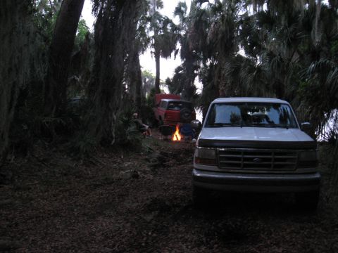 1994 Ford Bronco - A campsite scene with two trucks, a campfire, and people relaxing in a wooded area.