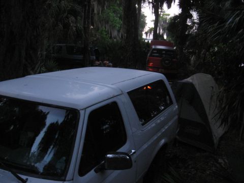 1994 Ford Bronco - A white SUV is parked next to a tent in a wooded area, with a red SUV visible behind it.