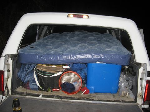 1994 Ford Bronco - The bed of a white pickup truck is filled with camping gear, including an air mattress, cooler, and buckets, ready for an outdoor excursion.