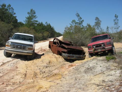 1994 Ford Bronco - Three trucks are stuck in the mud on a dirt road surrounded by trees.