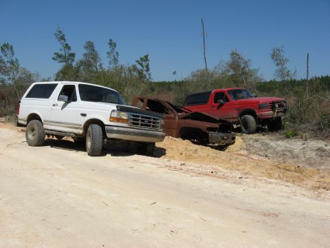 1994 Ford Bronco - Two Ford Broncos and a rusted vehicle chassis are parked on a dirt road with trees and brush in the background.
