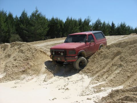 1994 Ford Bronco - A red 1994 Ford Bronco tackles a challenging off-road course with sandy inclines and clear blue skies.