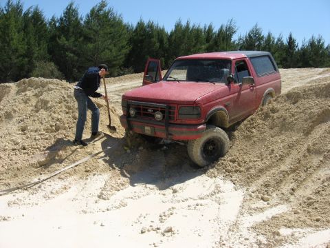 1994 Ford Bronco - A red Ford Bronco is stuck in a sandy area, with a person attempting to dig it out.