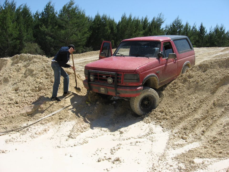 Red Ford Bronco stuck in sand, driver digging with shovel. Front grille and headlights visible