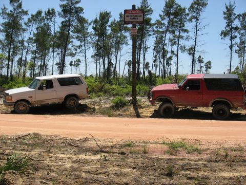 1994 Ford Bronco - Two Ford Broncos are parked on a dirt road next to a sign warning of unexploded bombs.