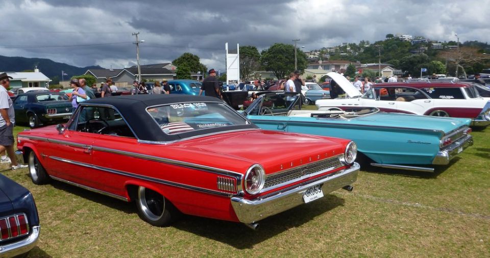 Red classic car with a black top parked on grass at an outdoor car show. Other classic cars are visible in the background