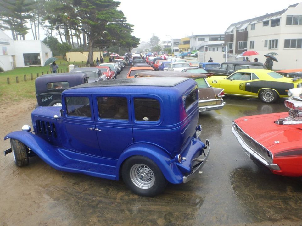 Blue hot rod sedan parked on wet pavement, other classic cars in background