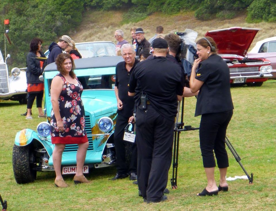 Woman in floral dress stands by a light blue hot rod with a film crew nearby. Other vintage cars are in the background