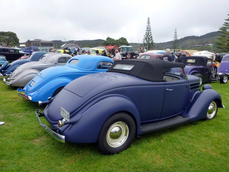 Dark blue convertible hot rod with black top, parked on grass with other classic cars
