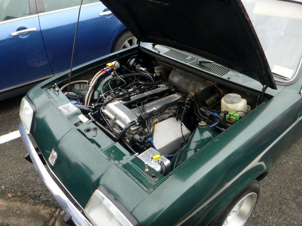 Green car engine bay with a V8 engine, chrome accents, and visible hoses. A blue car is parked beside it