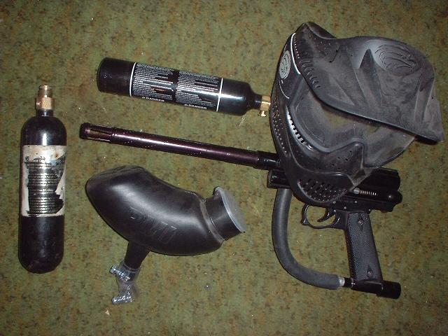 Black paintball marker, mask, hopper, and two gas tanks on a floor