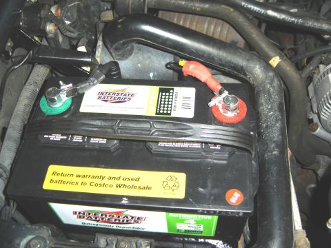 1996 Ford Bronco - Close-up of a car battery with visible brand and recycling labels, surrounded by engine components.