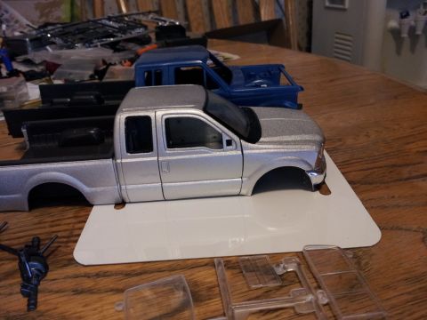 1995 Ford Bronco - Two model pickup trucks are shown on a wooden table, one silver and one blue, with various model parts scattered around.