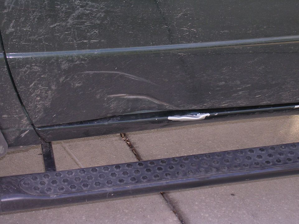 Side of a dark-colored SUV with a dent and scratches on the lower door panel, above a textured running board
