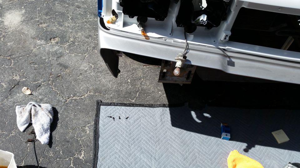 Car front end with bumper removed, showing wiring and light assembly. Tools and rags are on the ground