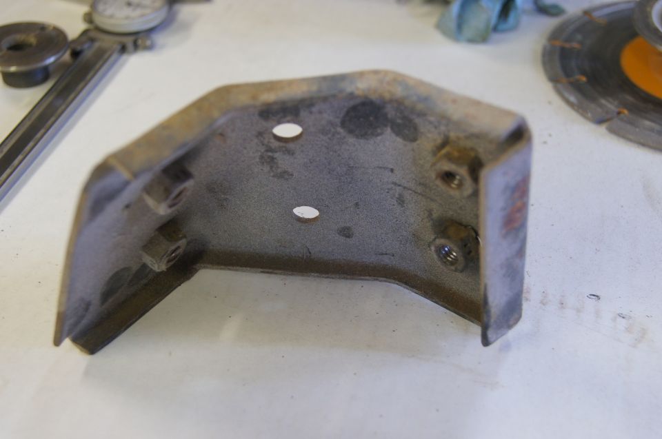 Rusty metal bracket with two bolt holes and nuts attached, on a white surface with tools