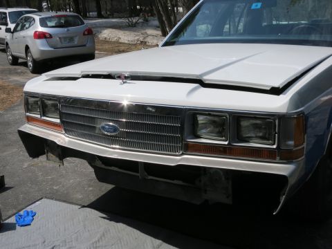 1991 Ford LTD Crown Victoria - The front of a white Ford Crown Victoria is shown with its hood open and bumper removed, revealing the grille and headlights.