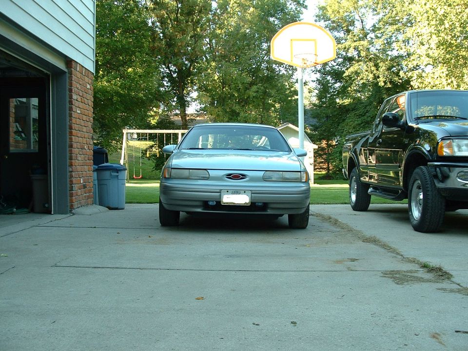 Front view of a silver-blue Ford Taurus sedan parked in a driveway next to a black pickup truck