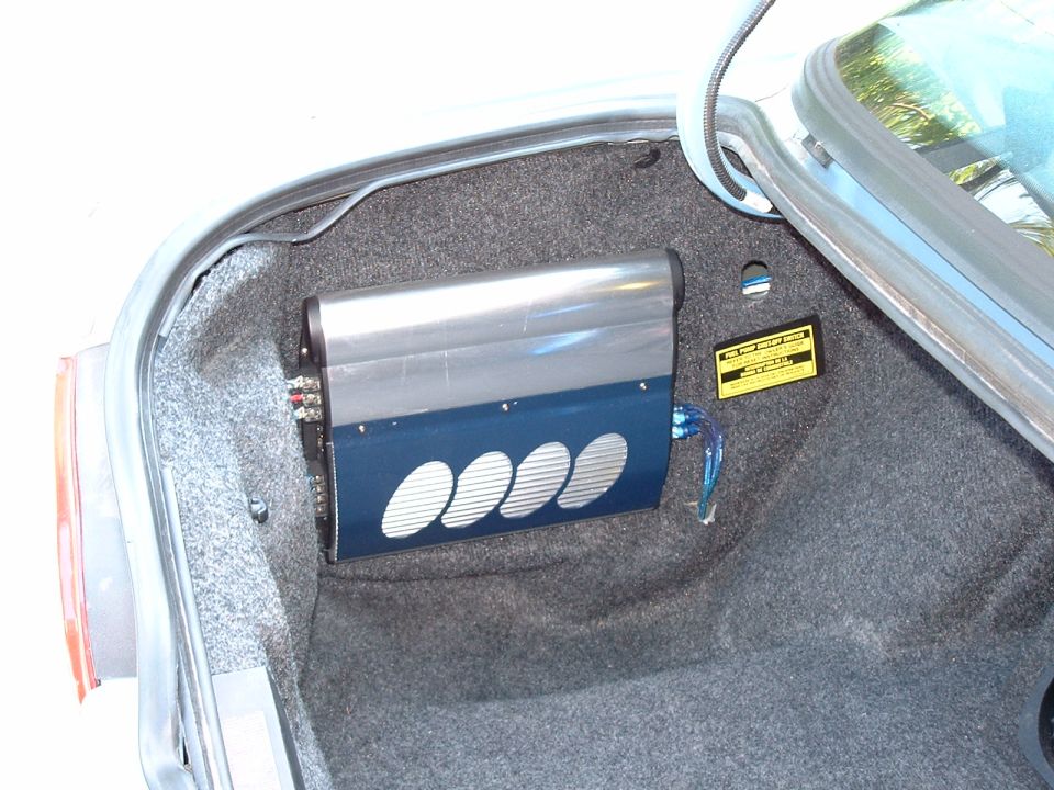 Blue and chrome car amplifier mounted in carpeted sedan trunk with warning label