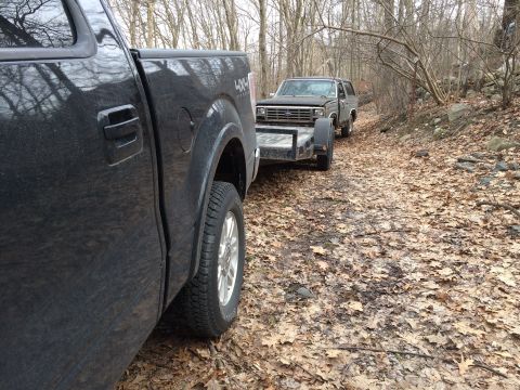 1986 Ford Bronco - A black Ford Bronco is being towed on a trailer through a wooded area by a pickup truck.
