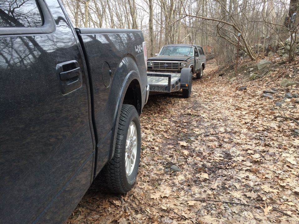 Black Ford Bronco on a trailer, towed by a black pickup truck on a dirt road