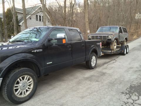 1986 Ford Bronco - A black Ford F-150 tows a vintage Ford Bronco on a trailer down a rural road.