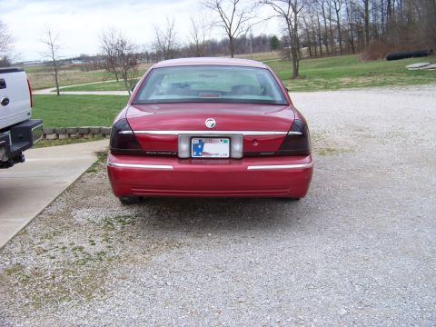1999 Mercury Grand Marquis - A red sedan is parked in a driveway next to a pickup truck, with trees and a field in the background.